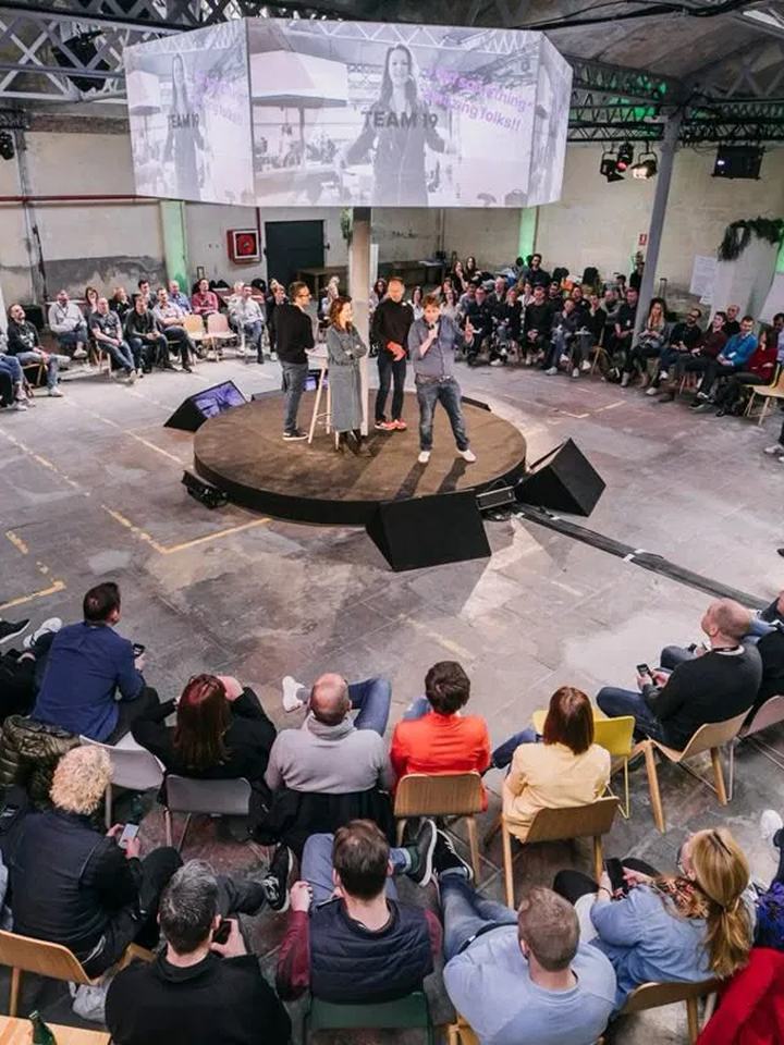 Plateia sentada em c&iacute;rculo ao redor de um palco redondo durante um encontro de comunidade em um galp&atilde;o