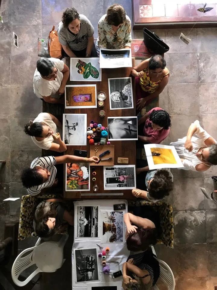 Vista de cima de um grupo reunido ao redor de uma mesa comprida, trabalhando com fotografias e materiais impressos